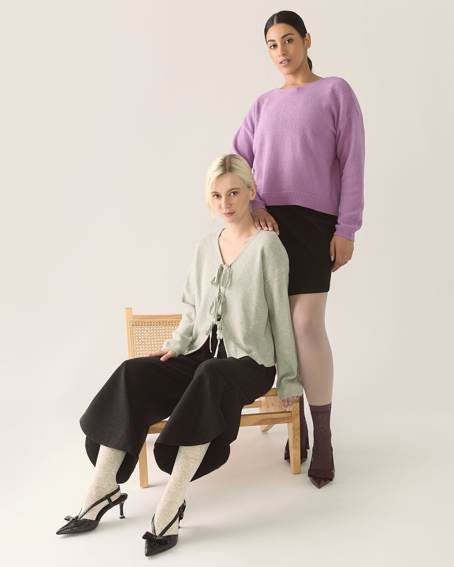 Two women posing against a white background, one sitting on a chair wearing a heather grey sweater and black pants, the other standing wearing a purple sweater and black skirt with tights.