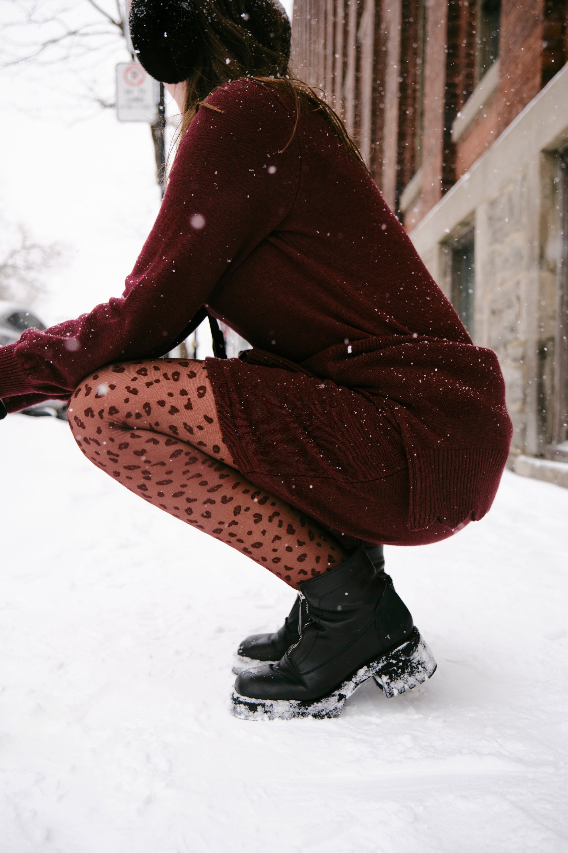 Person in a burgundy outfit with black shoes and patterned tights crouching in the snow.
